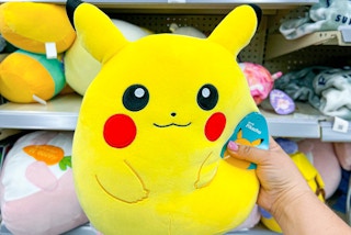 Someone holding a Pikachu Squishmallow in front of some shelves