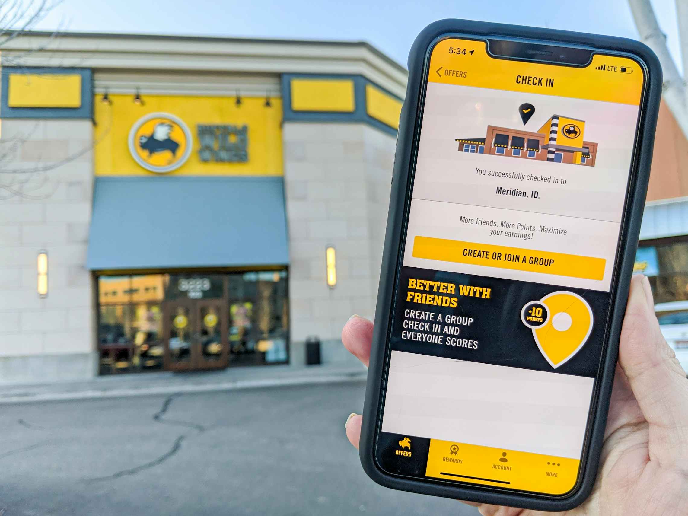 A person holding their iPhone up showing the check-in page of the Buffalo Wild Wings mobile app in front of a Buffalo Wild Wings storefro...