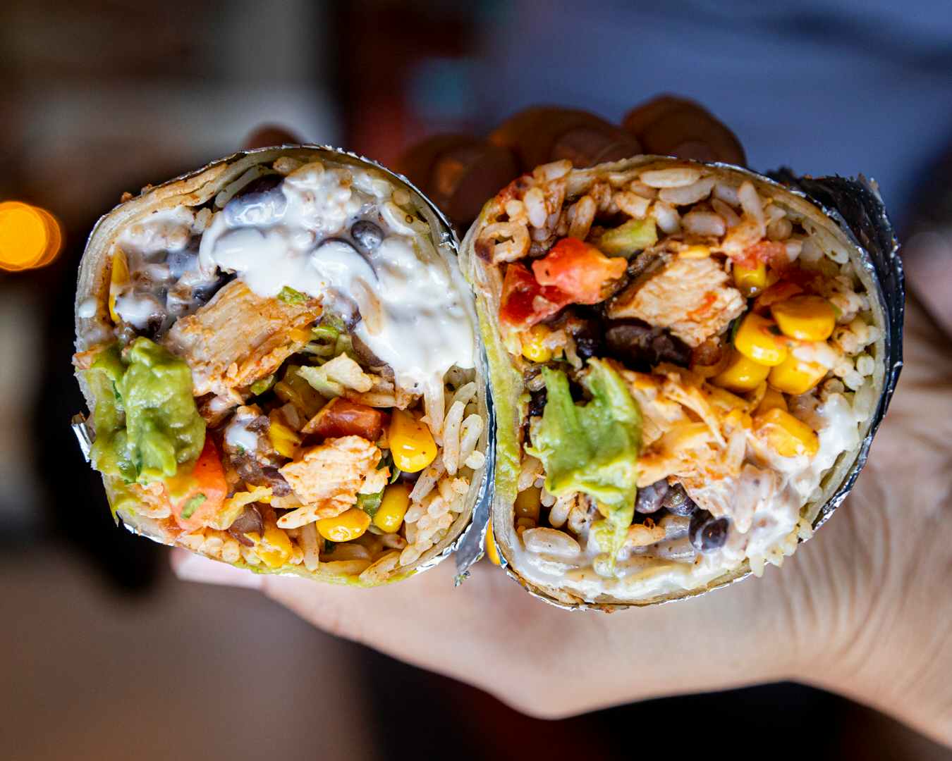 A person's hand holding up two halves of a burrito, showing the inside. The burrito is filled with meat, cheese, beans, rice, ...