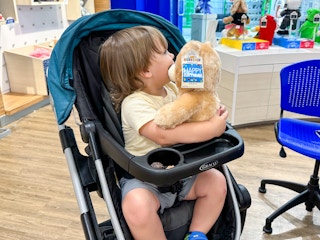 A little kid holding a Build-A-Bear Birthday Bear while sitting in a stroller