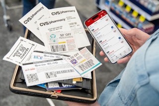 A person holding a cellphone displaying the CVS app next to a stack of coupons sitting on a wallet in the person's other hand.