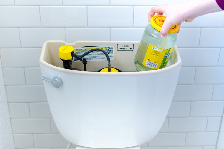 Person putting a jar filled with water into the tank of a toilet.