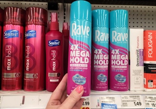 A person's hand holding up a bottle of Rave hair spray in front of a shelf at Kroger.