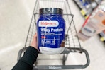 hand holding walgreens brand whey protein powder over shopping cart