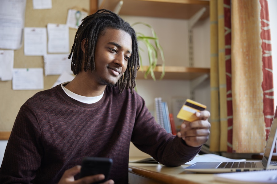 A college student sitting at a desk in a dorm room, looking at a credit card