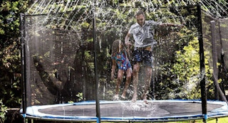 dad and daughter jumping on a trampoline with a sprinkler