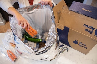 A person pulling carrots out of the cooler bag next to a blue apron meal kit box