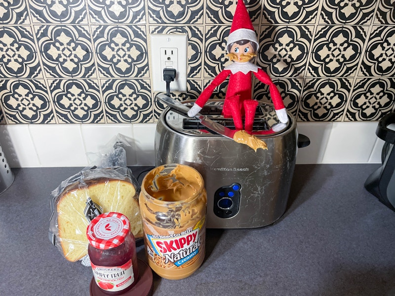 an elf on the shelf doll sitting on a toaster with a knife loaded with peanut butter and peanut butter on its face with bread, jam, and j...