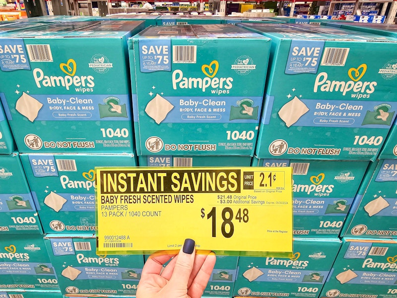person holding a sign for $18.48 sale price on pampers baby wipes in front of boxes of wipes