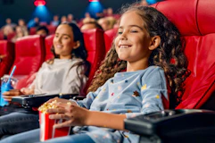 a girl holding a bag of popcorn watching a movie at the movies with her mom 