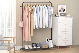 A rolling clothing rack stands between a window and a dresser against a wall, full of clothes and shoes