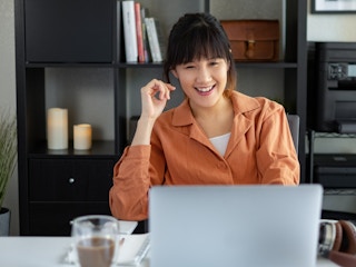 A woman sitting in a home office, smiling at a laptop.