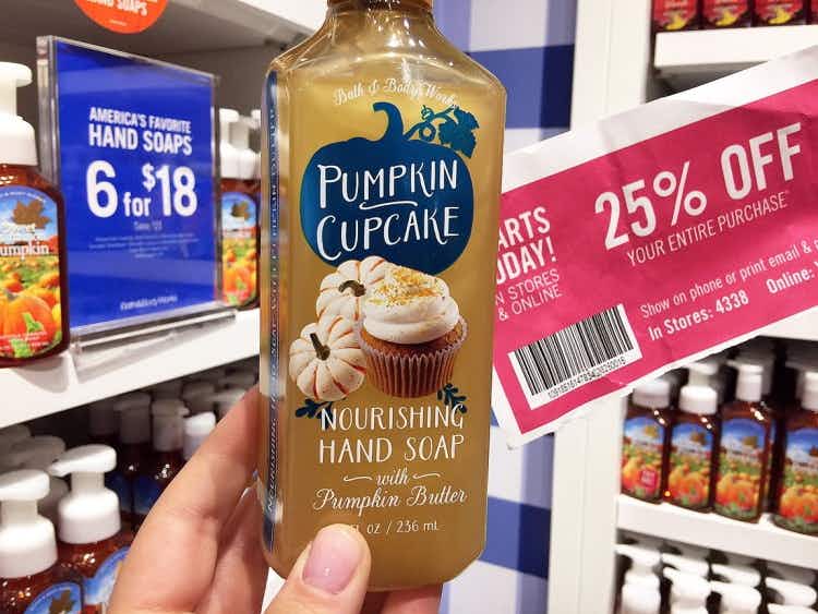 A person's hand holding up a Pumpkin Cupcake scented hand soap and a coupon for 25% off next to a sign that reads "hand soaps 6 for $18" ...