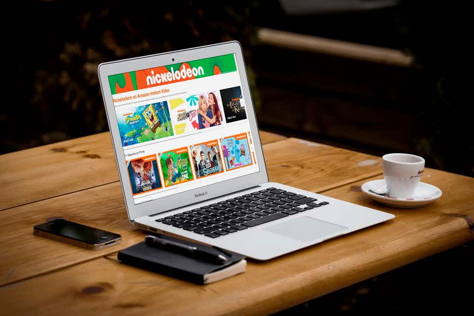 A laptop open on a table displaying a web page that features Nickelodeon shows. Sitting next to the laptop is a notebook and pen, a cellp...
