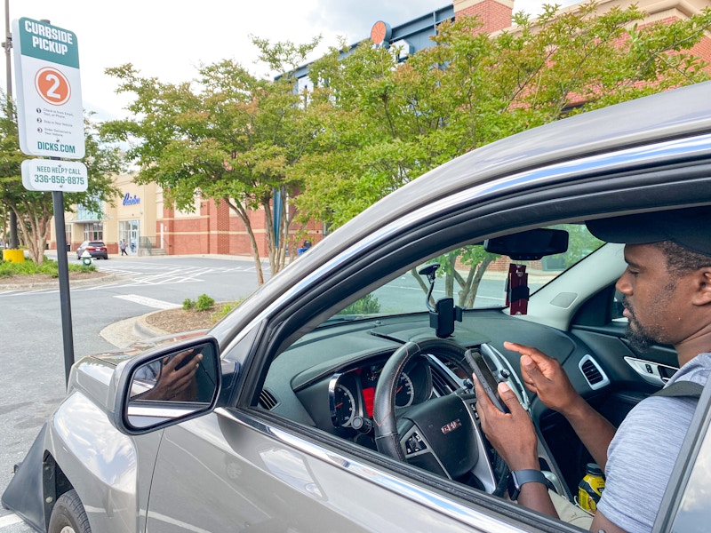 a man sitting in the car on his phone parked in curbside pickup outside of dicks sporting goods