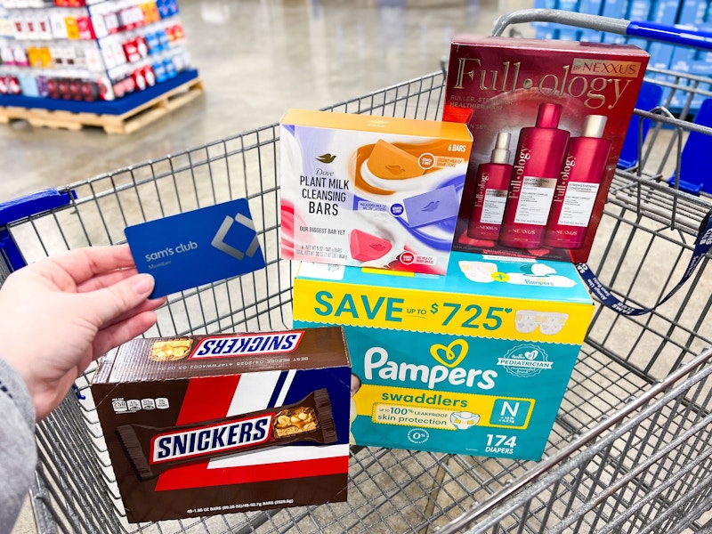 person holding a sams club membership card over a cart with snickers, dove soap, nexxus hair care, and pampers diapers