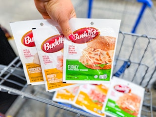 hand holding three packs of buddig lunch meat over three packs in walmart cart