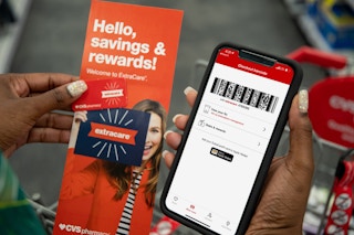 A woman holding an iPhone displaying her ExtraCare barcode on the CVS app, a rewards key chain card, and an extracare rewards pamphlet in...