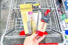 person holding loreal mascara, eyeliner, foundation, and lipstick over a cart