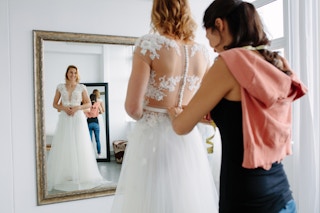 Reflection in mirror of female trying on wedding dress with women assisting her.