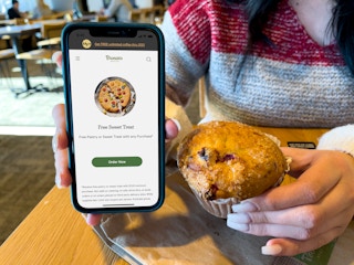 A person holding a cell phone displaying a free sweet treat next to a muffin at Panera.