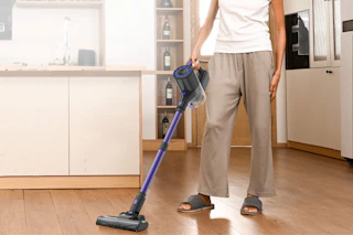 The bottom half of a person in a beige kitchen vacuuming the hardwood floor with a purple and grey cordless stick vacuum cleaner