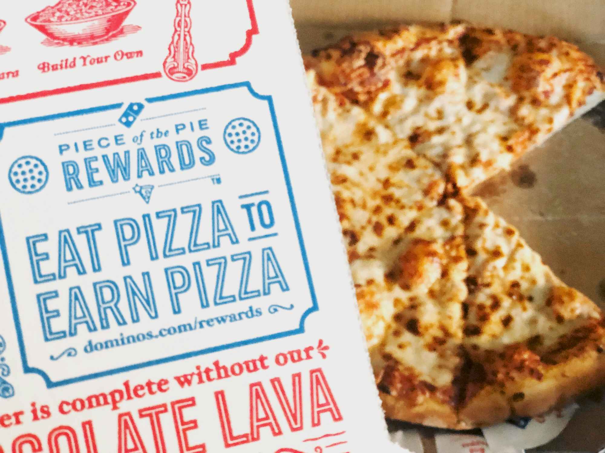 A Domino's pizza box advertising their Piece of the Pie Rewards program being held over a pizza with some slices taken from it.