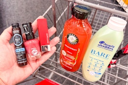 person holding hask hair oil, sally hansen nail polish, and a lipstick over a cart with herbal essences and head & shoulders shampoo in it