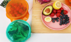 colorful stretch lids on bowls of food next to a plate of fruit on a cutting board