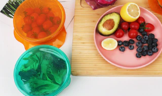 colorful stretch lids on bowls of food next to a plate of fruit on a cutting board