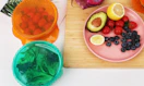 colorful stretch lids on bowls of food next to a plate of fruit on a cutting board