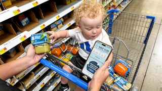 a person holding gerber baby food and holding a phone with gerber rewards on screen next to a baby in a walmart shopping cart