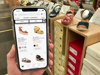 An iPhone displaying the DSW app showing clearance shoes, held next to similar shoes in the store.