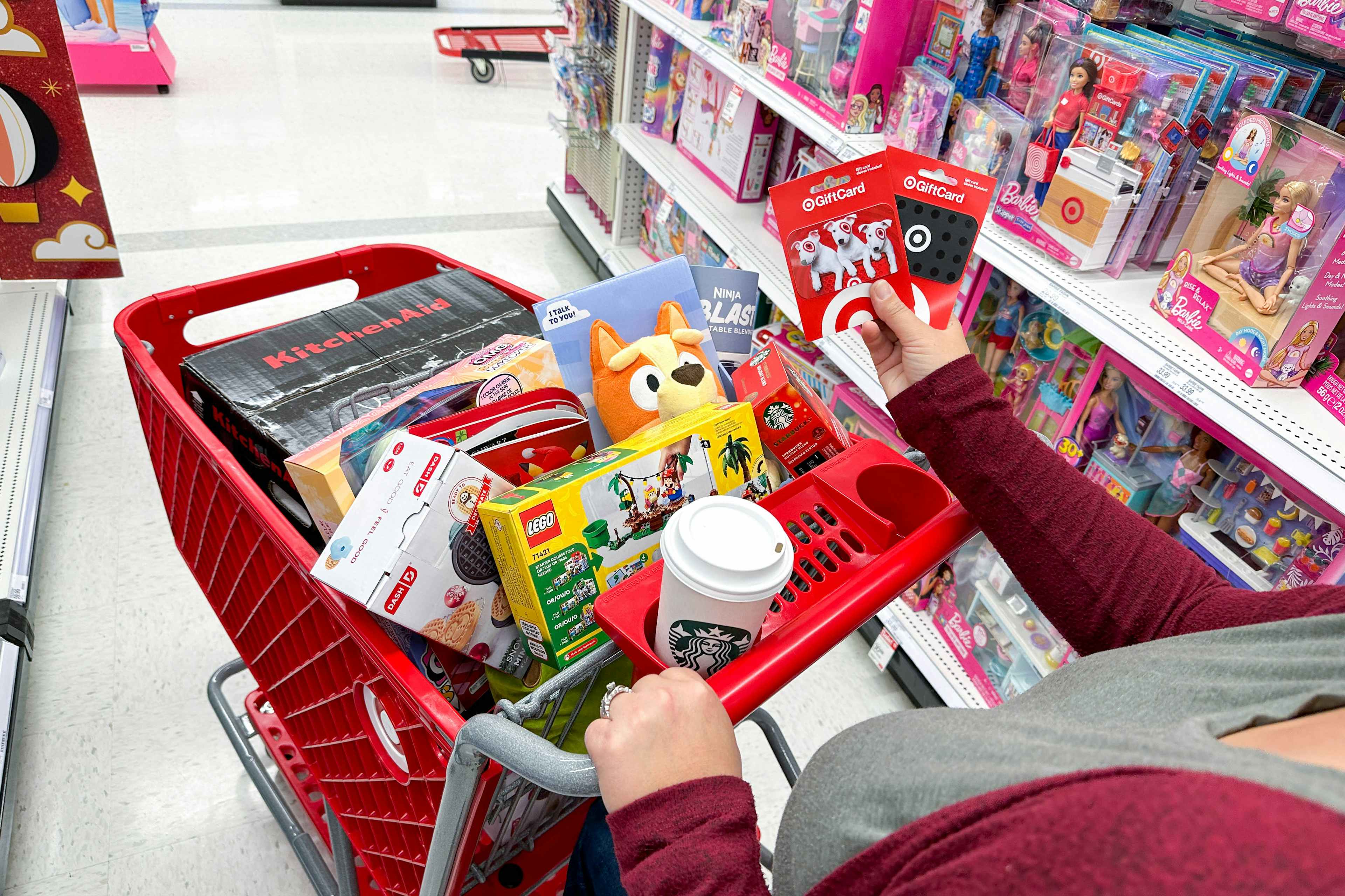 A woman pushing a target shopping cart filled with toys and holding two target gift cards in her right hand