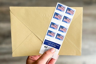 A person's hand holding up an envelope and some American flag Forever postage stamps.