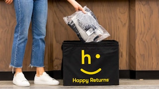 Person placing a a package of folded clothes into a black bin that shows happy returns logo in yellow
