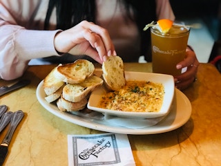 A person sitting at a table at The Cheesecake Factory, dipping a piece of bread into a bowl of dip and holding a drink with their other ...