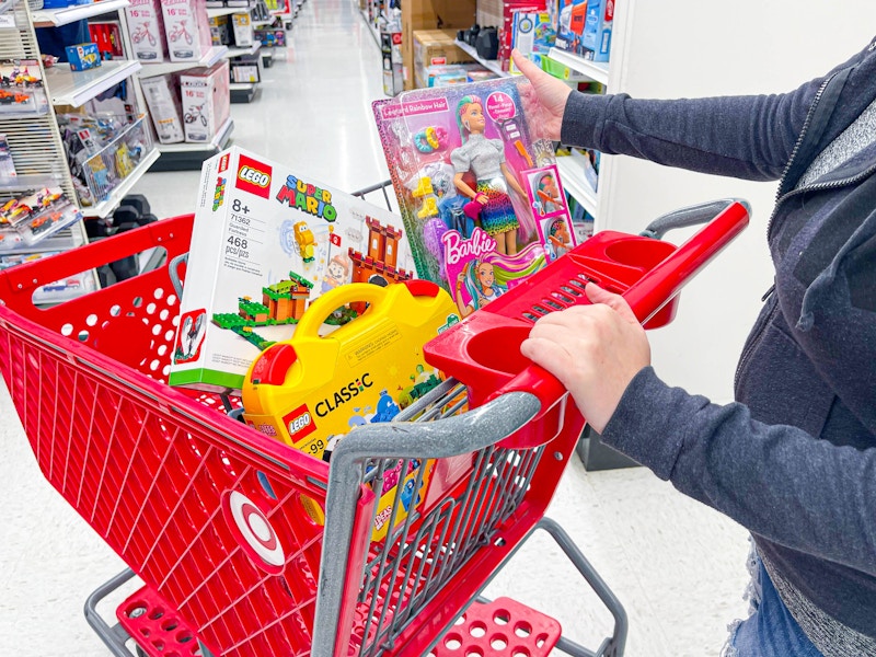 A person putting a Barbie doll into the basket of a Target shopping cart that has LEGO toys in it.