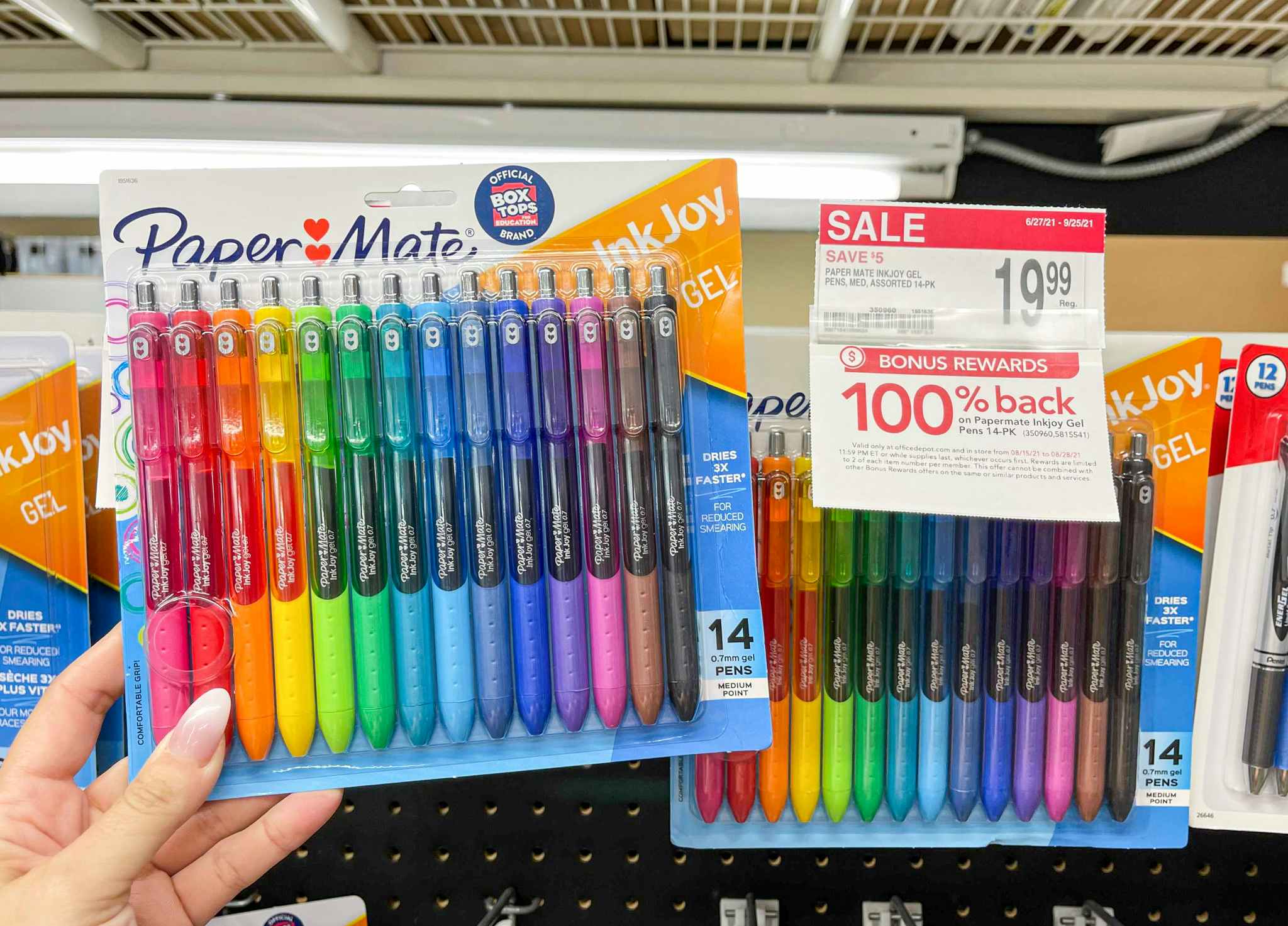 A person's hand holding a 14-pack of PaperMate gel pens in front of a display with more pens at Office Depot.
