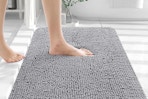 A foot steps on a plush chenille grey bath mat.