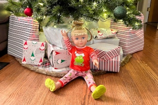  a walmart grinch doll sitting on the floor under a christmas tree next to gifts. 