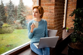 A person sitting by a window with a mug of coffee and a laptop on their lap.