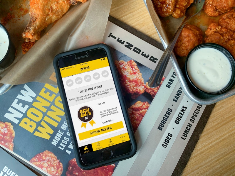 An iPhone sitting on top of a menu on a table, open to the Buffalo Wild Wings app's Offer page. The page shows a 25% off deal that can be...