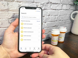 Someone holding a cell phone displaying the GoodRx app search screen next to some prescription medications on a table