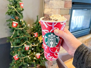 A person holding a Starbucks holiday drink cup in front of a little tree decorated with Starbucks ornaments