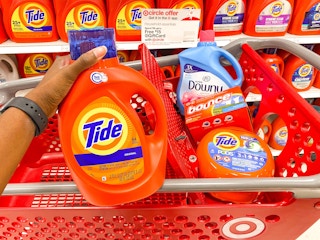 hand holding up a bottle of Tide in a Target aisle in front of a shopping cart that contains Tide pods, Downy liquid, and Bounce dryer ...
