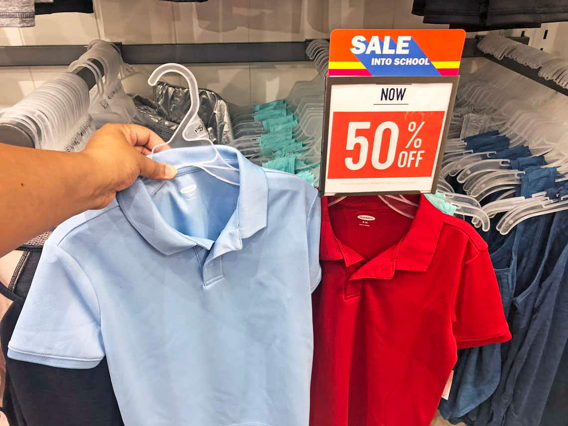 A person's hand holding up a child's school uniform polo shirt on next to another shirt hanging below a sign that reads, "Sale into ...