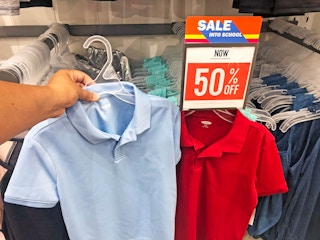 A person's hand holding up a child's school uniform polo shirt on next to another shirt hanging below a sign that reads, "Sale into ...