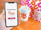 A person holding a cell phone displaying the intro page for the new Dunkin Rewards app next to a coffee and food bag on a table.