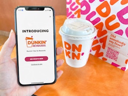 A person holding a cell phone displaying the intro page for the new Dunkin Rewards app next to a coffee and food bag on a table.